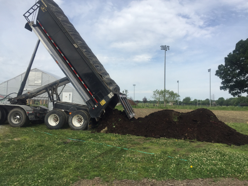 dump truck unloading leaf mold compost onto field with greenhouse and tall light poles in background