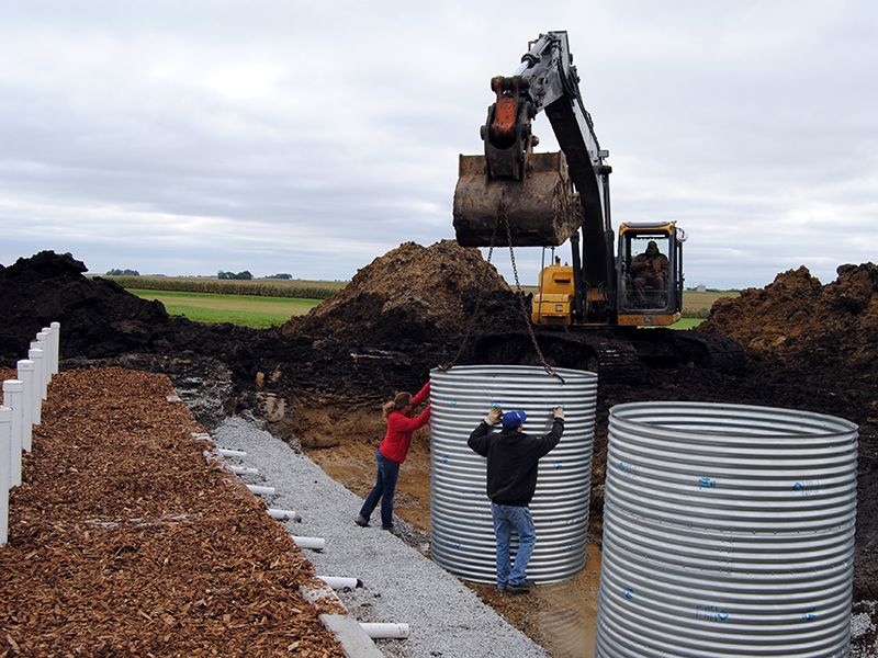 An excavator installs culverts in a field area with people standing by