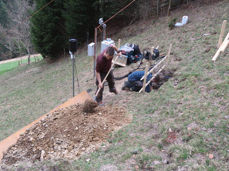 person shoveling dirt and rocks on wooden board on side of hill with various equipment in background